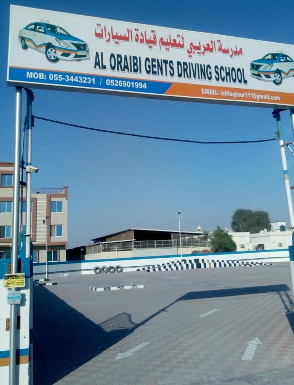 Entrance signboard of Al Oraibi Gents Driving School, featuring Arabic and English text along with car illustrations. The driving school premises are visible in the background, including a practice area with traffic markings and a nearby building under a clear sky.