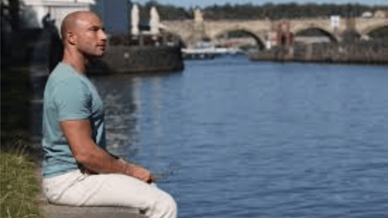 Pascal Bachmann sitting by the water, reflecting with a scenic bridge in the background, embodying mindfulness and success.